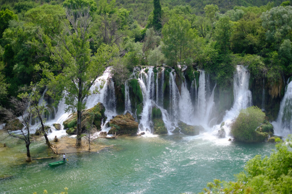 A panoramic view of the Kravica Waterfalls, showing multiple streams of water cascading over a lush, green semicircular cliff into a clear lake.